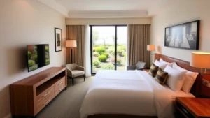 Modern hotel room interior with clean white bedding, wooden desk, flat-screen TV, and natural light from window overlooking landscaped grounds with ornamental shrubs and flowers