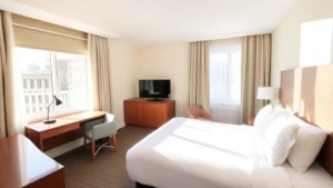 Bright, sunny hotel room with modern furnishings, comfortable bed with fresh linens, work desk, and windows showing natural light streaming across contemporary bedroom furniture